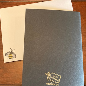 K&ouml;nnte beinhalten: Eine schwarze Karte mit einer goldenen Bienenillustration und dem Text "HAND STAMPED" und "STAMPIN' UP!" in einem goldenen Rechteck.