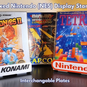 May include: Three boxed Nintendo Entertainment System (NES) video games on display stands. The games are The Goonies II, Bionic Commando, and Tetris. The stands are white with interchangeable plates that feature the game's logo.