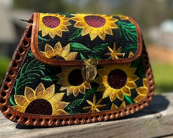 May include: A black crossbody bag with embroidered sunflowers in yellow, green, and burgundy. The bag features a brown leather trim and a gold-colored clasp. The bag's design is a floral theme.