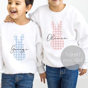Op de afbeelding: Twee witte sweatshirts met een gingham konijn ontwerp. Het konijn aan de linkerkant is blauw en zegt "George" en het konijn aan de rechterkant is roze en zegt "Olivia". T-shirt beschikbaar.