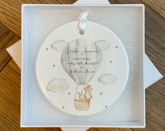 May include: A round, white ceramic ornament with a hot air balloon design. The balloon is gray, and the basket holds a rabbit and a bear. The ornament includes the text "Noah James, Christened Sunday 26th April 2023 at St Michael's Church."