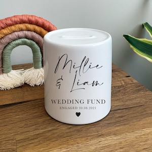 May include: White ceramic wedding money box with black lettering. The box says "Millie & Liam Wedding Fund Engaged 30.08.2021" with a small heart symbol.