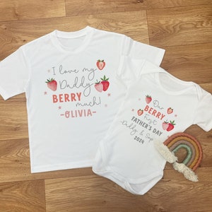 May include: Two white t-shirts with a strawberry theme. The larger shirt says "I love my Daddy BERRY much! -OLIVIA-." The smaller shirt says "Our First BERRY Father's Day -Daddy & Sop 2024."