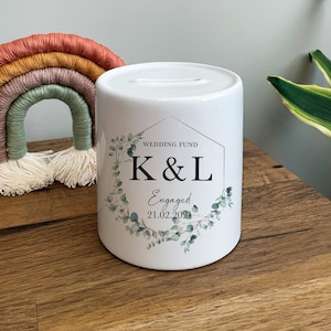May include: White ceramic money jar with a gold geometric frame and eucalyptus leaves. The jar has the text "Wedding Fund K&L Engaged 21.02.2021" printed on it.