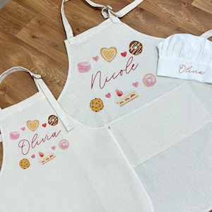 Puede incluir: Dos delantales blancos con diseños de pastelería rosa y marrón. El delantal más grande tiene el nombre "Nicole" bordado. El delantal más pequeño tiene el nombre "Olivia" bordado. Un gorro de chef blanco con el nombre "Olivia" bordado.