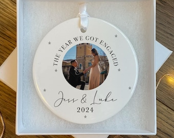 May include: White ceramic ornament with a photo of a couple getting engaged. The ornament says "The year we got engaged" with the couple's names "Jess & Luke" and the year "2024".
