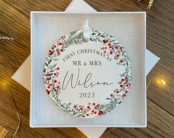 Op de afbeelding: Witte kerstbal van keramiek met een groen en rood bloemig kransontwerp. De kerstbal heeft de tekst "First Christmas as Mr. & Mrs. Wilson 2023".