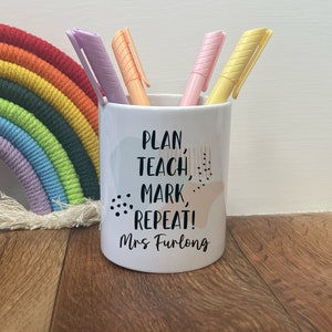 Puede incluir: Portalápices de cerámica blanca con el texto "PLAN, TEACH, MARK, REPEAT! Mrs Furlong" y formas abstractas en colores pastel. El portalápices contiene cuatro resaltadores en morado, naranja, rosa y amarillo. Un arcoíris de macramé decora el fondo.