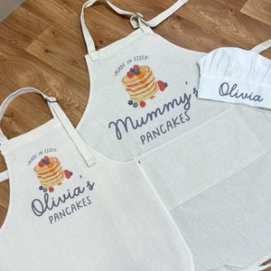 Puede incluir: Juego de delantales y gorro de chef de color blanquecino. Los delantales presentan un diseño de panqueques y los textos "Mummy's Pancakes" y "Olivia's Pancakes". El gorro de chef es blanco y lleva el nombre "Olivia".