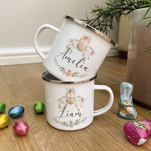 Può includere: Due tazze di smalto bianco con un design di coniglio e uovo. Le tazze sono personalizzate con i nomi "Amelia" e "Liam".