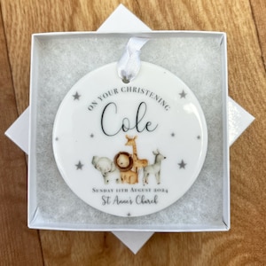 Puede incluir: Adorno redondo de cer&aacute;mica blanca con una cinta gris. El adorno tiene un patr&oacute;n de estrellas grises y una imagen de un le&oacute;n, una jirafa, un elefante y una cebra. El texto en el adorno dice "On your christening Cole Sunday 11th August 2024 St Anne's Church".