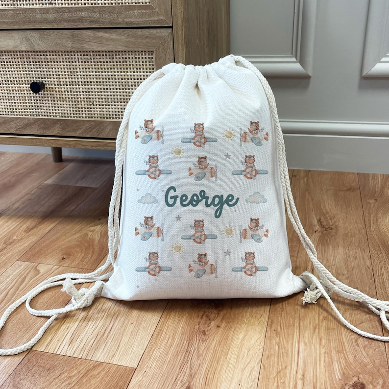 May include: A white canvas drawstring bag with a personalized name "George" printed on it. The bag features a repeating pattern of cute cartoon bears flying airplanes with green, blue, and yellow accents.