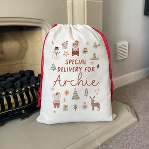 May include: White Christmas gift bag with red drawstring and a festive design featuring a snowman, a house, a bear, a deer, and a Christmas tree. The bag has the text "Special Delivery for Archie" printed on it.