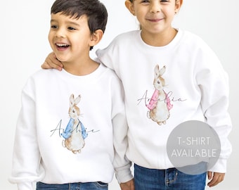 May include: Two white sweatshirts with Peter Rabbit illustrations. One sweatshirt has a blue jacket on the rabbit and the name "Archie" printed on it. The other sweatshirt has a pink jacket on the rabbit and the name "Amelia" printed on it. T-shirt available.