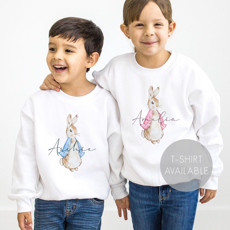 Puede incluir: Dos sudaderas blancas con ilustraciones de Peter Rabbit. Una sudadera tiene una chaqueta azul en el conejo y el nombre "Archie" impreso en ella. La otra sudadera tiene una chaqueta rosa en el conejo y el nombre "Amelia" impreso en ella. Camiseta disponible.
