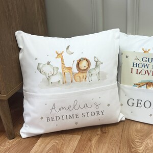 May include: White pillow with a pocket for a book. The pillow has a design of a giraffe, elephant, lion, and zebra with crowns on their heads. The text "Amelia's Bedtime Story" is printed on the pillow.