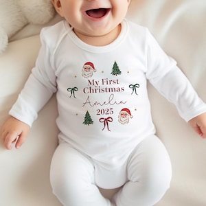 May include: White long-sleeved baby onesie with a Christmas theme. The onesie features the text "My First Christmas Amelia 2025" in burgundy script, along with illustrations of Santa Claus, Christmas trees, and bows.