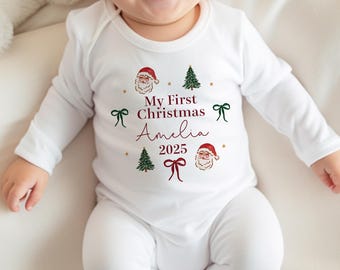 May include: White long-sleeved baby onesie with a Christmas theme. The onesie features the text "My First Christmas Amelia 2025" in burgundy script, along with illustrations of Santa Claus, Christmas trees, and bows.