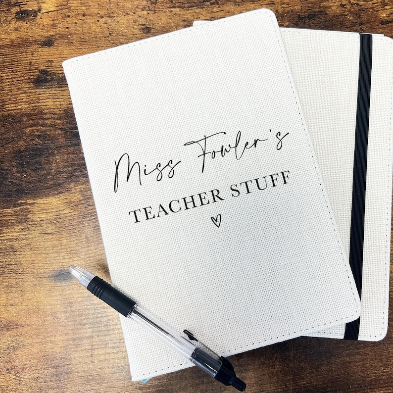 May include: Two cream-coloured notebooks with the text "Miss Fowler's TEACHER STUFF" in black, and a heart symbol. A black and clear pen lies on the notebook. One notebook has a black elastic band.