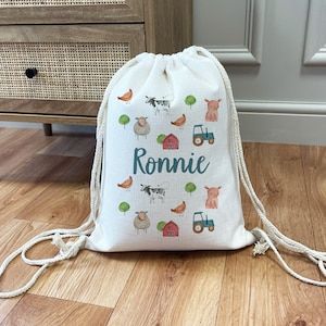 May include: A white drawstring bag with a farm animal theme. The bag features cows, pigs, sheep, chickens, and a tractor. The name "Ronnie" is printed in teal on the front of the bag.