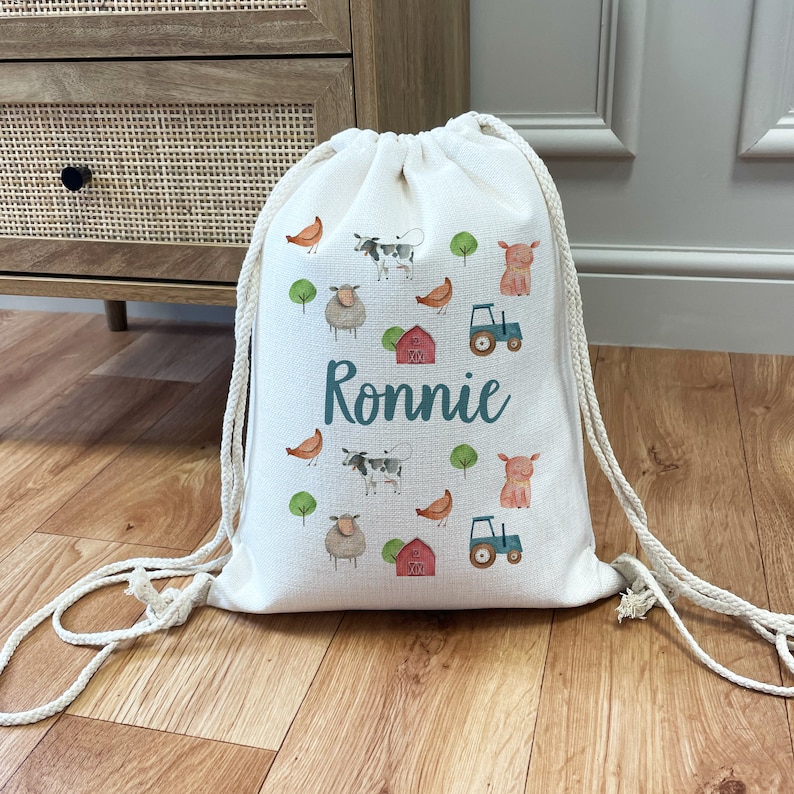 May include: A white drawstring bag with a farm animal theme. The bag features cows, pigs, sheep, chickens, and a tractor. The name "Ronnie" is printed in teal on the front of the bag.