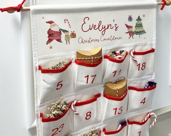 Puede incluir: Calendario de cuenta atrás navideño con bolsillos numerados y ribeteados en rojo. Presenta ilustraciones de Papá Noel, un elfo y un árbol de Navidad. El nombre "Evelyn's" está impreso encima de los bolsillos.