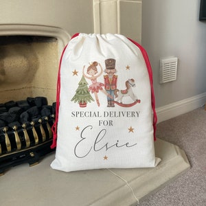 May include: White Christmas gift bag with red drawstring, featuring a Nutcracker, a ballerina, a rocking horse, and a Christmas tree. The bag has the text "Special Delivery for Elsie" printed on it.