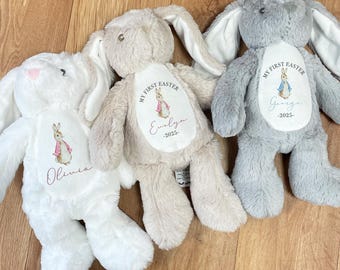 May include: Three plush bunny rabbits with embroidered details. Each bunny has a personalized patch with the text "My First Easter" and a name. The bunnies are white, light brown, and gray.