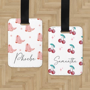 May include: Two white luggage tags with black straps. One tag has a pink and white cowboy boot pattern with the name "Phoebe" written in black. The other tag has a red and white cherry pattern with the name "Samantha" written in black.