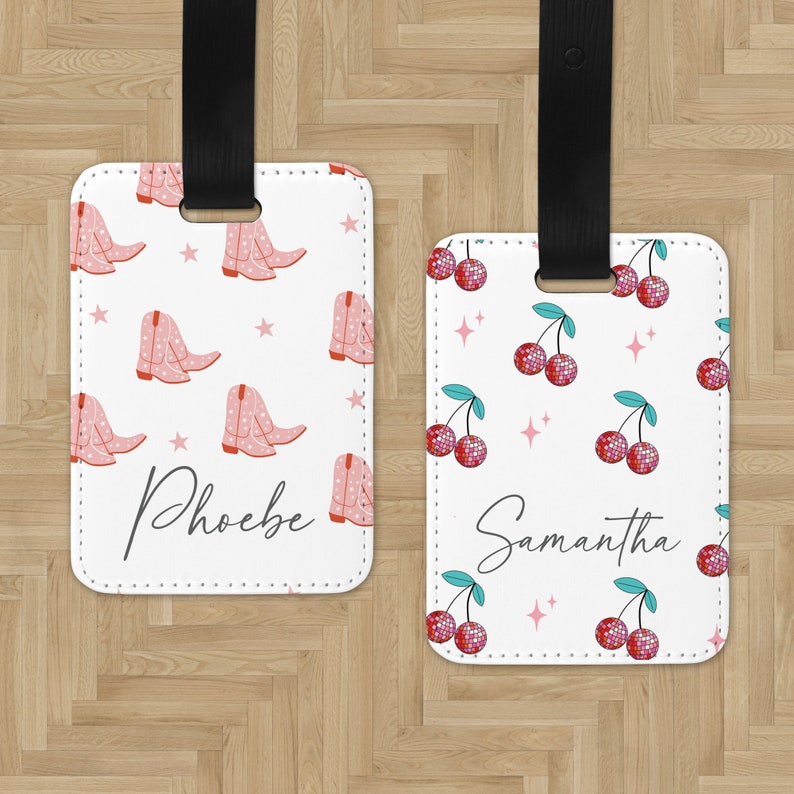 May include: Two white luggage tags with black straps. One tag has a pink and white cowboy boot pattern with the name "Phoebe" written in black. The other tag has a red and white cherry pattern with the name "Samantha" written in black.