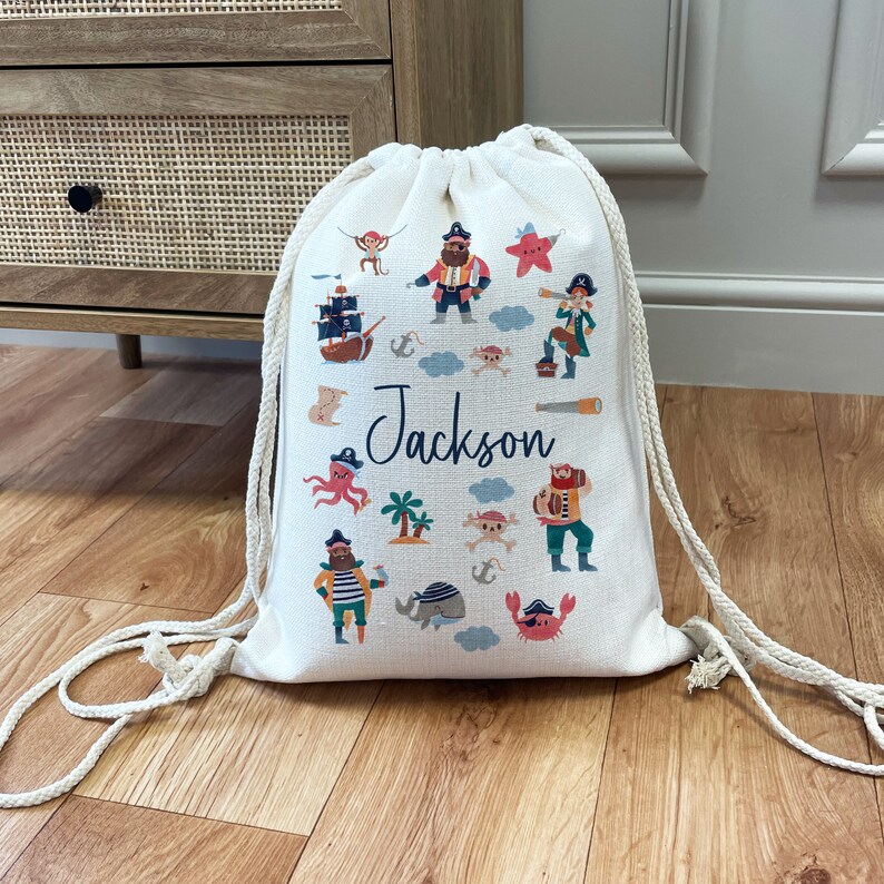 May include: A white drawstring bag with a pirate theme. The bag features a variety of colorful illustrations, including pirates, ships, and sea creatures. The name "Jackson" is printed in black on the front of the bag.