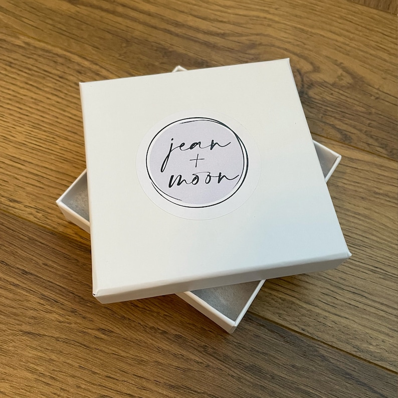 Puede incluir: Caja cuadrada blanca con una pegatina redonda que dice "jean + moon" en escritura cursiva negra.