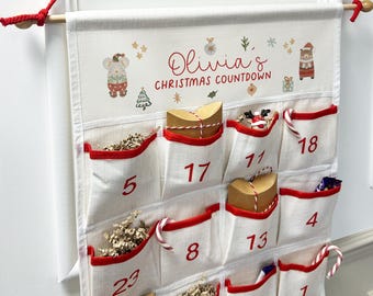 Puede incluir: Un calendario de cuenta regresiva navideño color crema con ribetes y bolsillos rojos. El calendario tiene el nombre "Olivia's" y el texto "Christmas Countdown" en escritura roja. Presenta ilustraciones festivas y bolsillos numerados.
