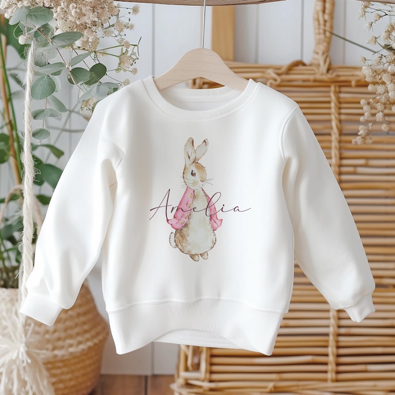 Puede incluir: Sudadera blanca con una ilustraci&oacute;n de Peter Rabbit y el nombre Amelia impreso en cursiva.
