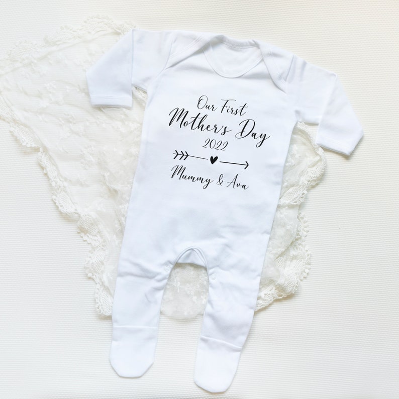 May include: White baby onesie with black text that reads "Our First Mother's Day 2022" with an arrow pointing to the right and the text "Mummy & Ava" below.