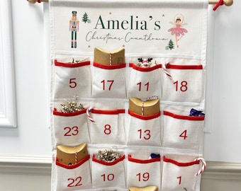May include: A fabric Christmas advent calendar with pockets, numbered 1-23, trimmed in red. The top features the text "Amelia's Christmas Countdown" with a nutcracker and ballerina illustration. The pockets are filled with small gifts.