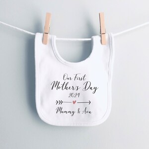 May include: White baby bib with black text that reads "Our First Mother's Day 2024" with a pink heart and arrow. The text "Mummy & Ava" is below the arrow.
