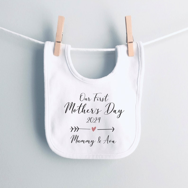 May include: White baby bib with black text that reads "Our First Mother's Day 2024" with a pink heart and arrow. The text "Mummy & Ava" is below the arrow.