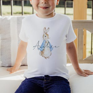 Puede incluir: Camiseta blanca con una ilustraci&oacute;n de Peter Rabbit con una chaqueta azul. El nombre "Archie" est&aacute; escrito en cursiva debajo del conejo.