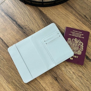 May include: A light blue passport holder with a pocket for a passport or credit card. The holder is lying on a wooden surface next to a burgundy passport.