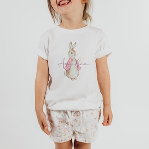 Puede incluir: Camiseta blanca con una ilustraci&oacute;n de acuarela de un conejo marr&oacute;n con una chaqueta rosa. El nombre "Amelia" est&aacute; escrito en cursiva debajo del conejo.