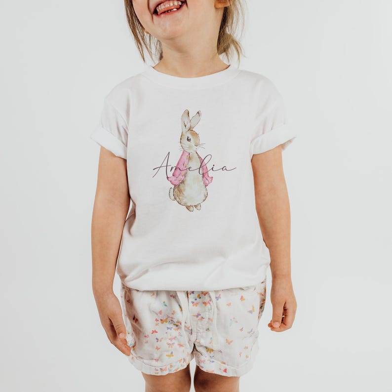Puede incluir: Camiseta blanca con una ilustraci&oacute;n de acuarela de un conejo marr&oacute;n con una chaqueta rosa. El nombre "Amelia" est&aacute; escrito en cursiva debajo del conejo.