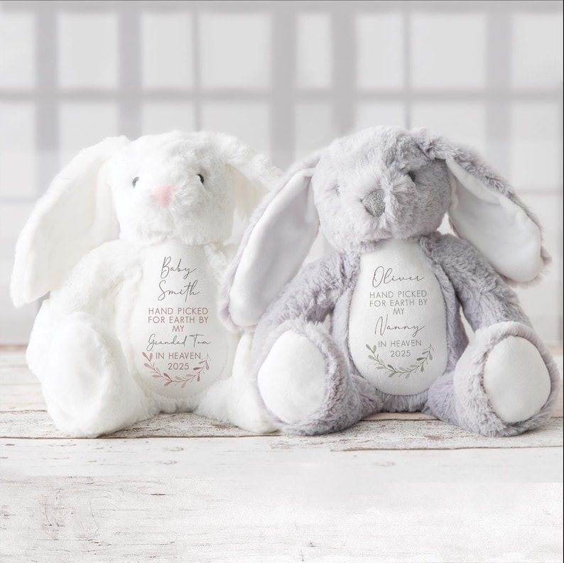 May include: Two plush bunny rabbits with embroidered messages. The white bunny has "Baby Smith HAND PICKED FOR EARTH BY MY Guanded Tra IN HEAVEN 2025" embroidered on it. The gray bunny has "Olivia HAND PICKED FOR EARTH BY MY Nanny IN HEAVEN 2025" embroidered on it.