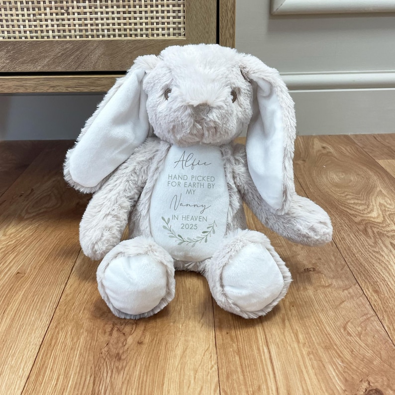 May include: A beige plush bunny rabbit with white ears. The bunny has embroidered text that reads "Alfie Hand Picked For Earth By My Nanny In Heaven 2023".