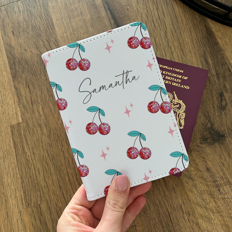 May include: White passport holder with a cherry pattern. The cherries are red and have a sparkly texture. The passport holder has the name Samantha written on it.