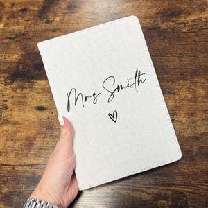 May include: A white notebook with the words "Mrs Smith" in black script and a heart symbol. The notebook has a textured surface and is held against a brown wooden background.