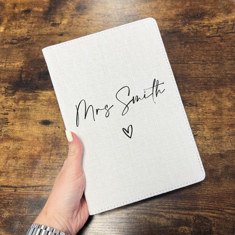 May include: A white notebook with the words "Mrs Smith" in black script and a heart symbol. The notebook has a textured surface and is held against a brown wooden background.