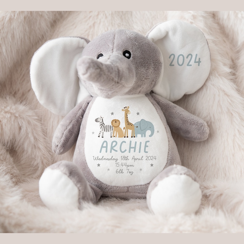 May include: A gray plush elephant stuffed animal with a white patch on its chest. The patch features a zebra, lion, giraffe, and elephant illustration, along with the name "Archie", the date "Wednesday 18th April 2024", the time "15:44pm", and the text "6lb 7oz". The elephant's ear has the year "2024" embroidered on it.