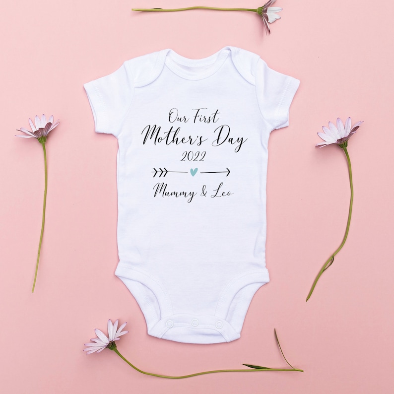 May include: White baby onesie with the text "Our First Mother's Day 2022" and an arrow with a heart pointing to the text "Mummy & Leo" on a pink background.