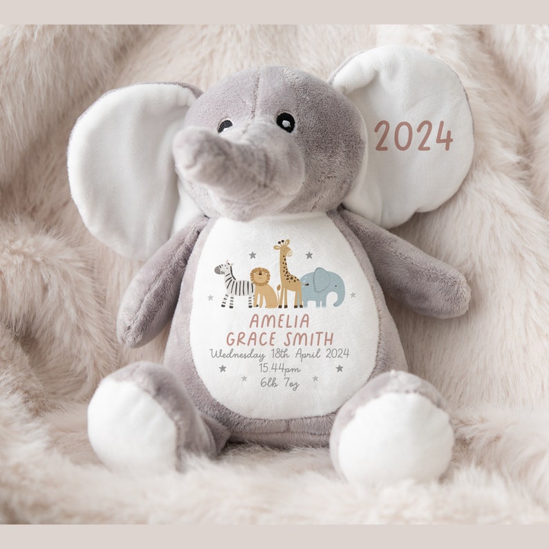 May include: Gray plush elephant stuffed animal with a white patch on its chest. The patch has a design of a zebra, lion, giraffe, and elephant with stars. The text on the patch reads "Amelia Grace Smith, Wednesday 18th April 2024, 15.44pm, 6lb 7oz". The elephant's ear has the text "2024" in pink.